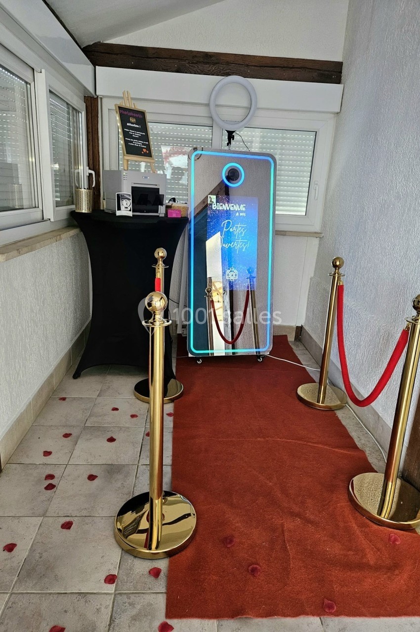 Miroir interactif lumineux entouré de poteaux dorés et tapis rouge, dans une pièce lumineuse avec décoration sobre.