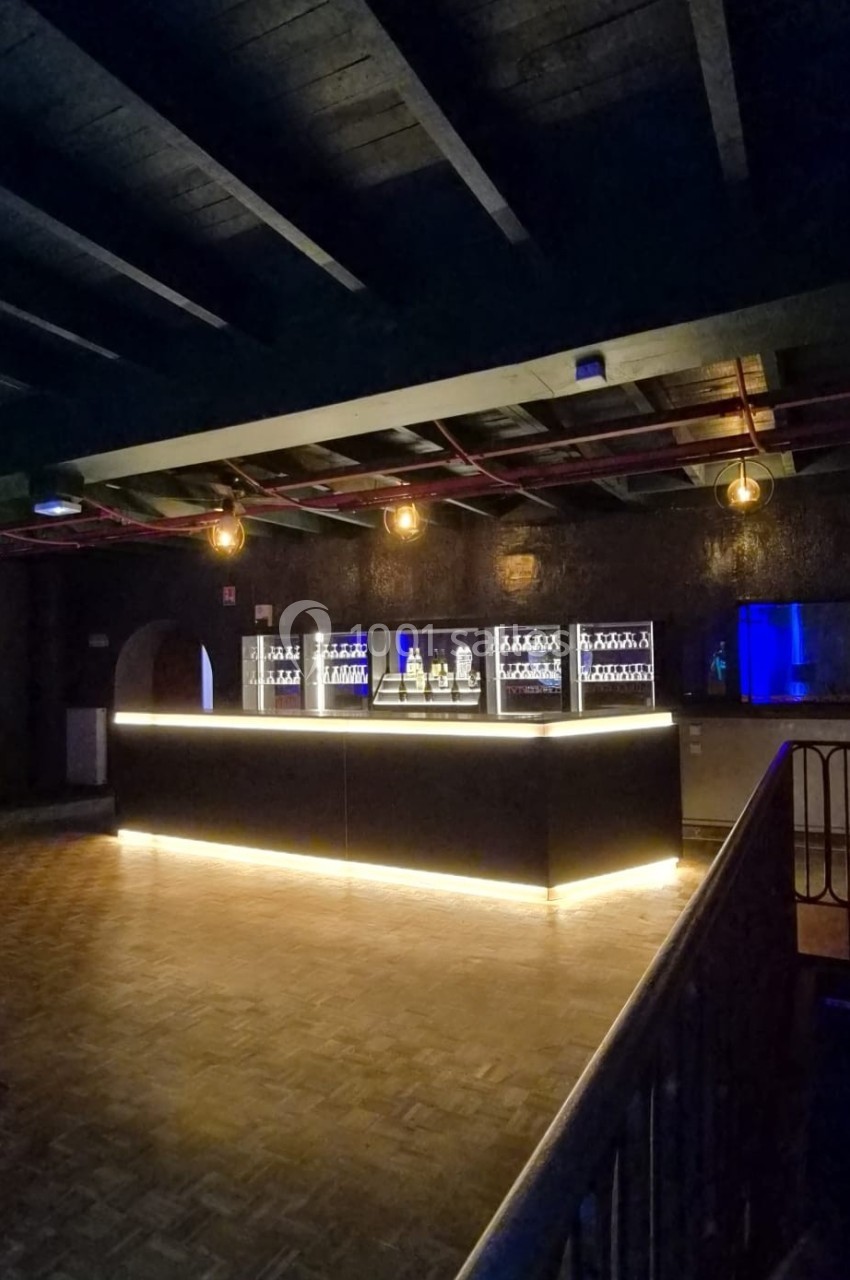 Bar moderne éclairé avec des étagères de bouteilles, situé dans une grande salle sombre au parquet en bois.