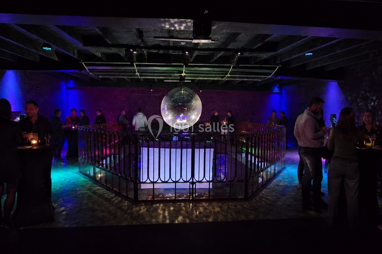 Salle sombre avec une piste de danse centrale, éclairée par une boule à facettes et des lumières colorées, entourée de…