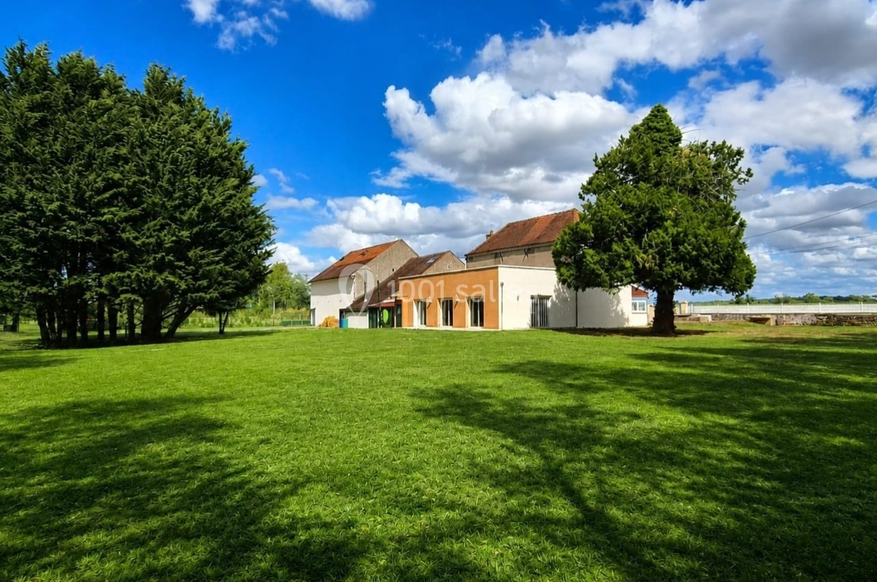 Grande maison avec toit en tuiles entourée d'arbres, située dans un vaste jardin sous un ciel bleu parsemé de nuages.