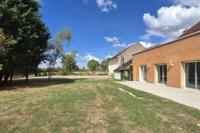 Un jardin avec pelouse, arbres et un bâtiment beige à gauche sous un ciel partiellement nuageux.