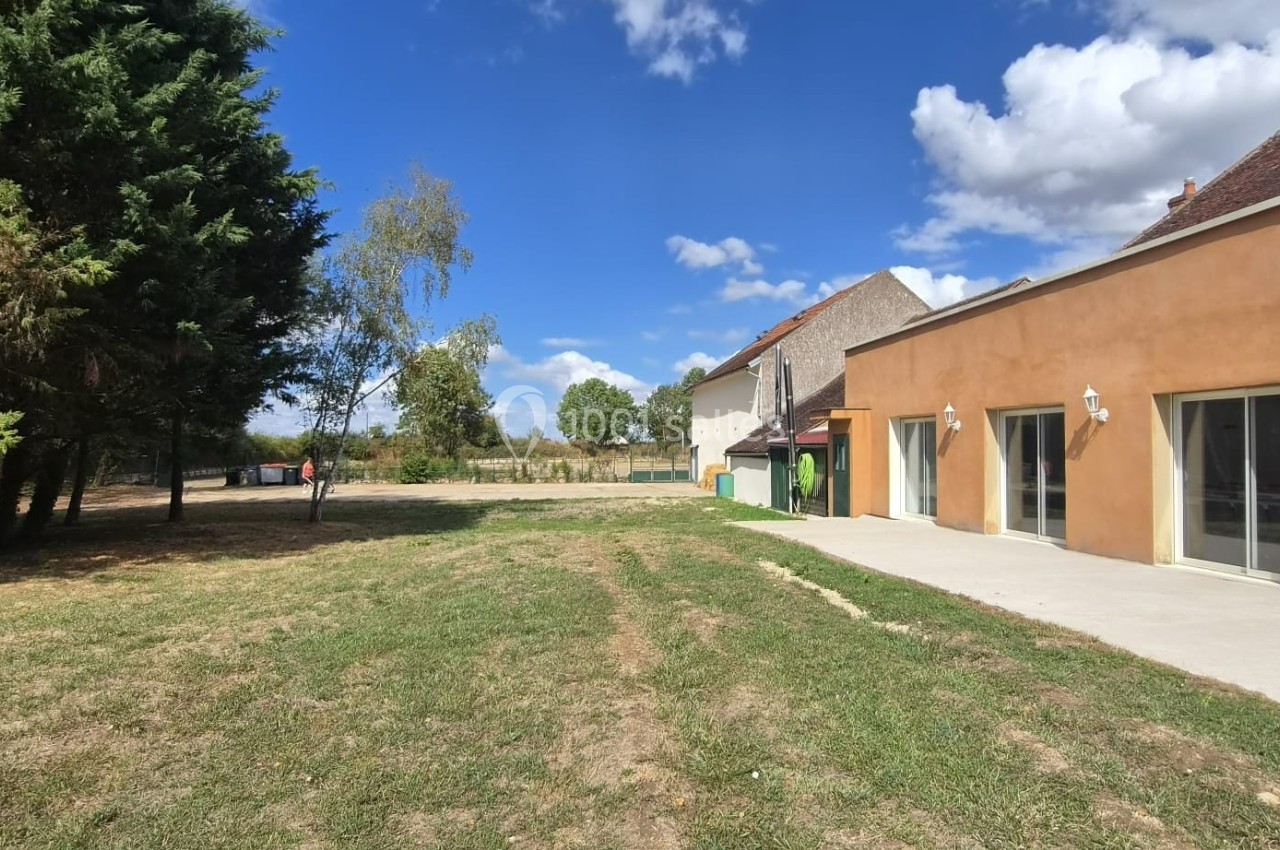 Jardin avec pelouse bordé d'arbres, adjacent à une maison aux murs beige et grandes baies vitrées, sous un ciel bleu.