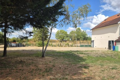 Un jardin avec pelouse, arbres et un bâtiment beige à gauche sous un ciel partiellement nuageux.