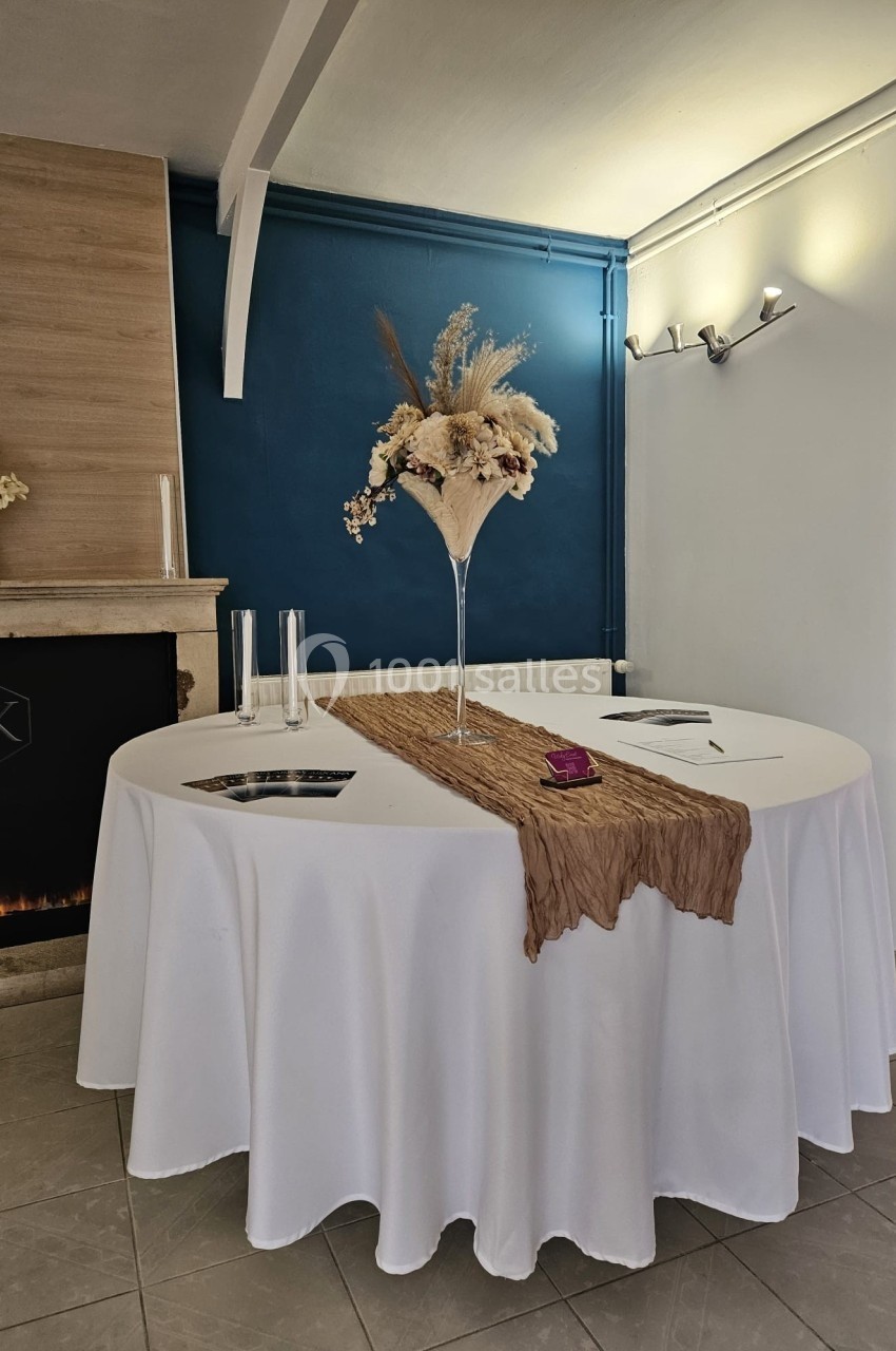 Table ronde décorée d'une nappe blanche, d'un chemin de table beige et d'un vase fleuri, dans une pièce élégante.