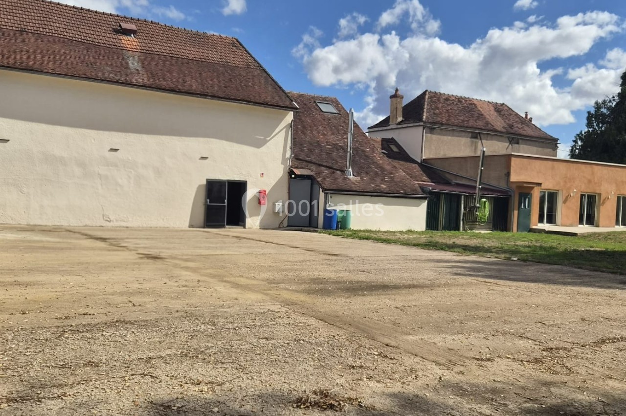 Bâtiments ruraux rénovés avec façades claires, entourés d'une cour en gravier sous un ciel partiellement nuageux.