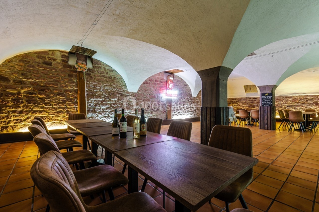 Salle voûtée en pierre avec table en bois, chaises rembourrées et bouteilles de vin, éclairage chaleureux.