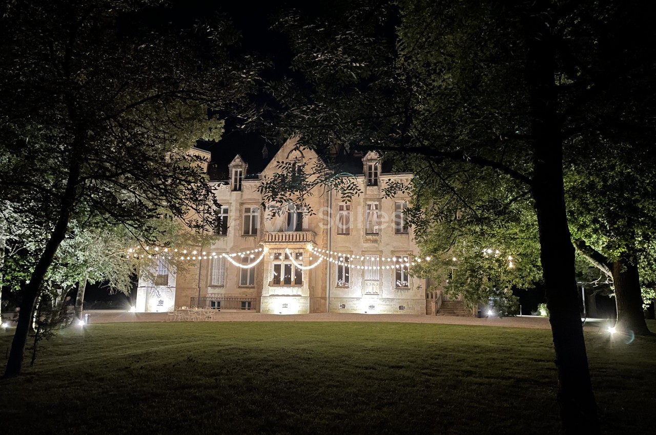 Château de pierre claire illuminé la nuit, façade ornée de guirlandes lumineuses, entouré d'arbres et pelouse.