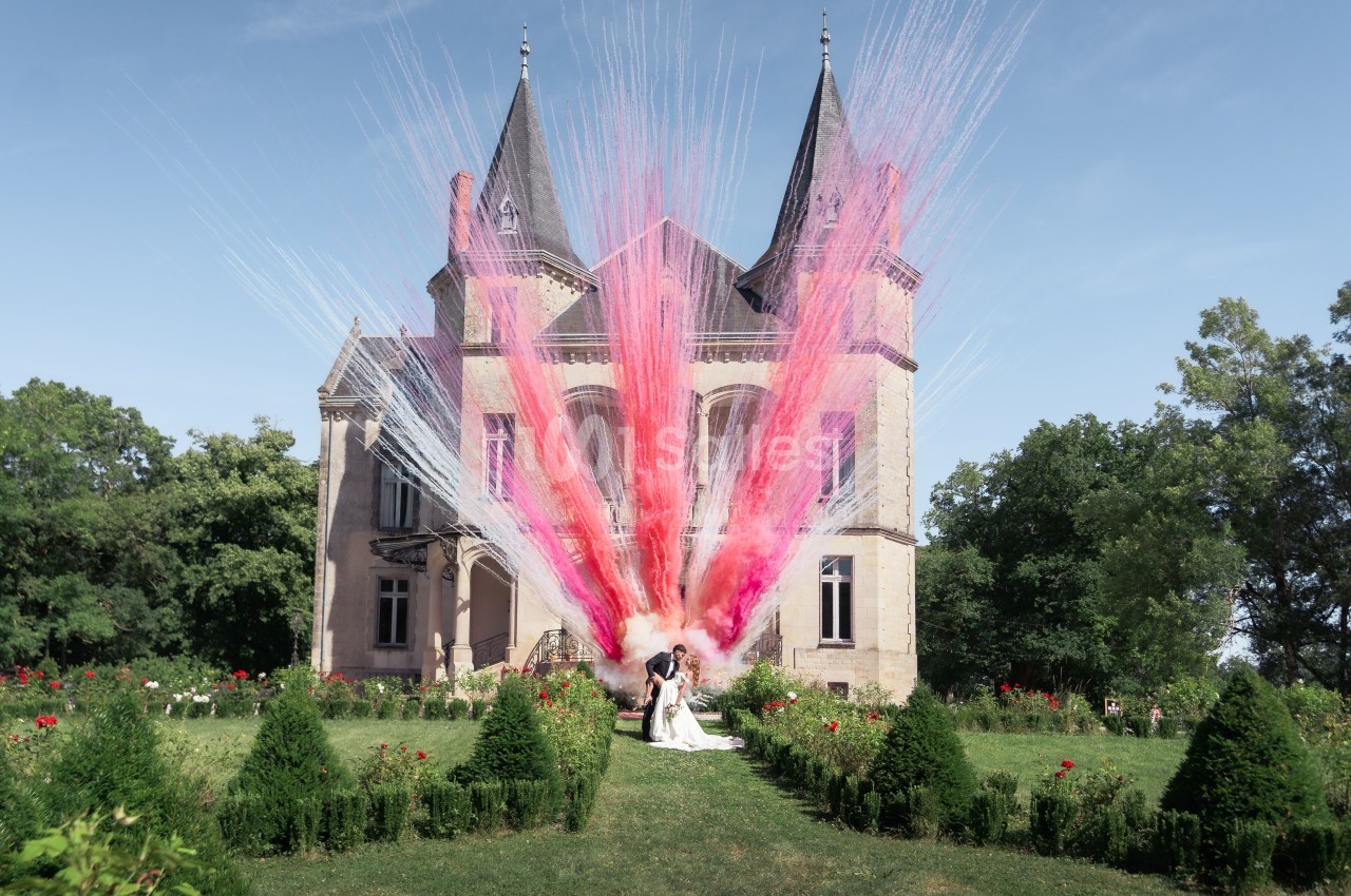 Château de pierre avec deux tours pointues entouré de verdure, poudre de couleur rose et blanche explosant devant la façade, mariés au premier plan.