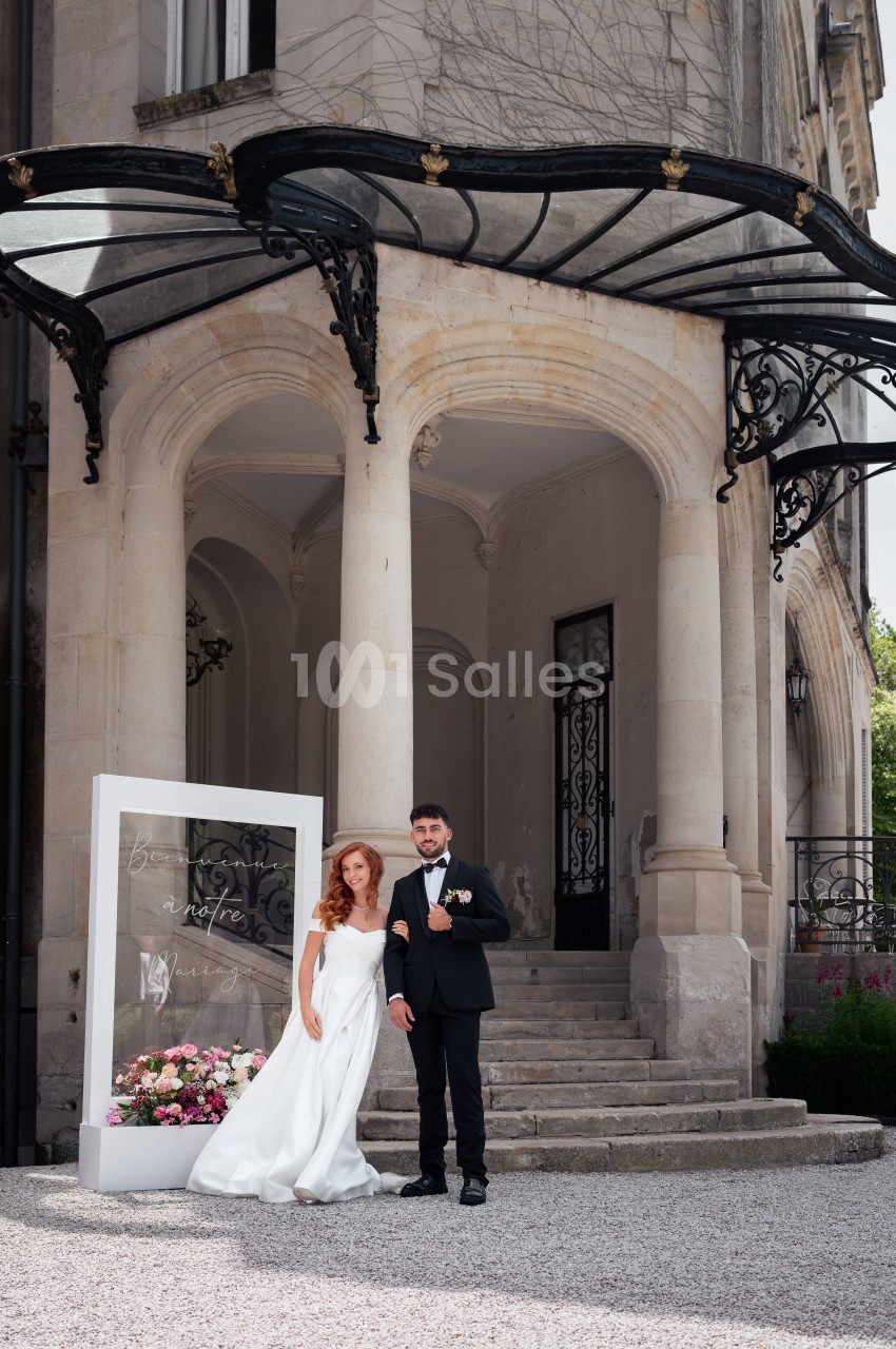 Couple de mariés devant la façade en pierre du château avec auvent noir et colonnes classiques.