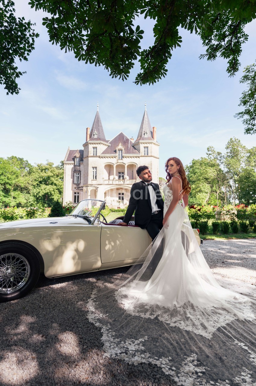 Mariés posant devant une voiture décapotable beige, château aux deux tourelles en arrière-plan.