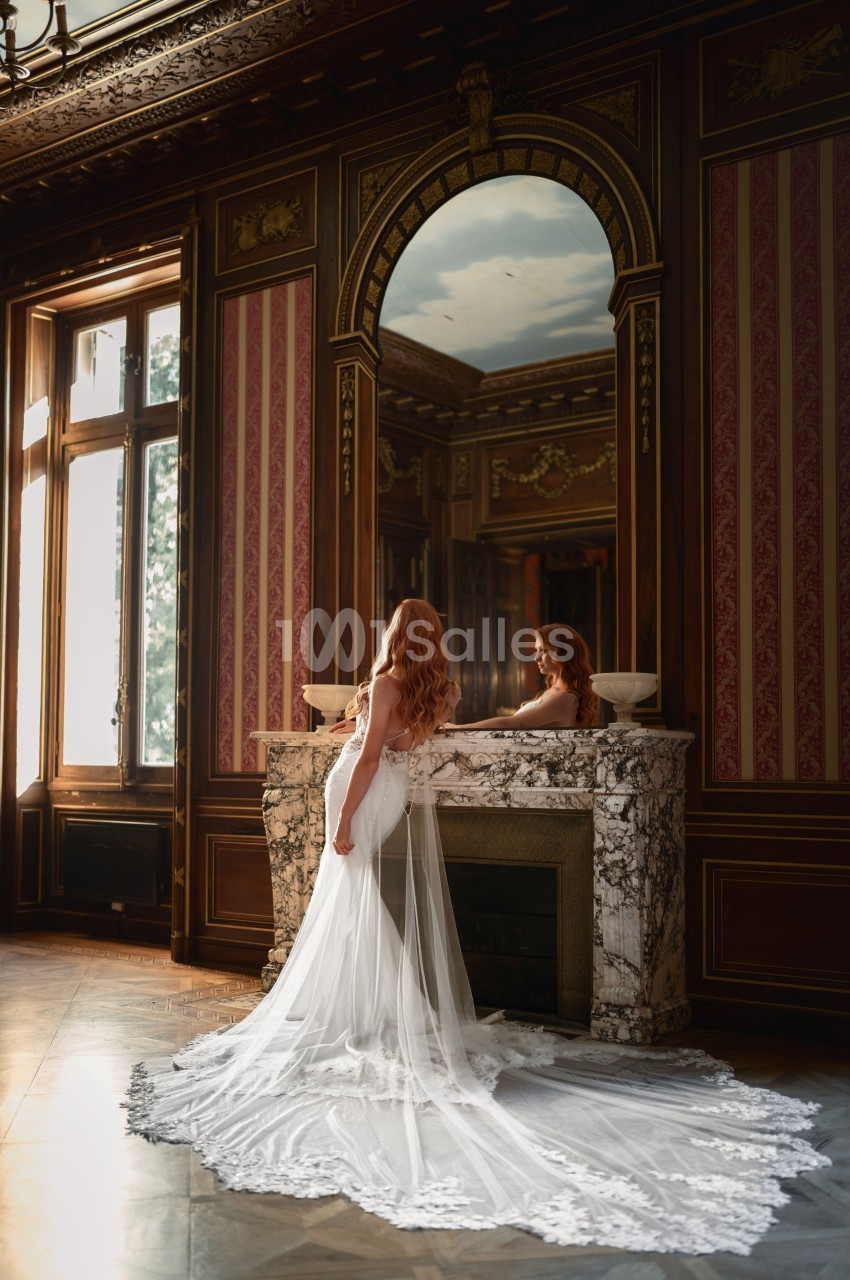 Mariée en robe blanche devant cheminée en marbre dans salon aux boiseries dorées et murs rouge bordeaux.