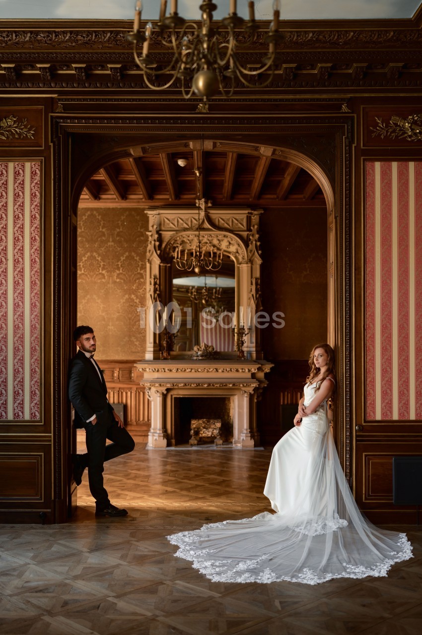 Salle classique avec cheminée ornementée, plafond à caissons et boiseries, deux personnes en tenue de cérémonie.