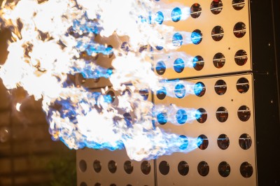 Un homme de dos observe un mur de dispositifs avec des flammes jaillissant à l'extérieur, entouré de végétation.