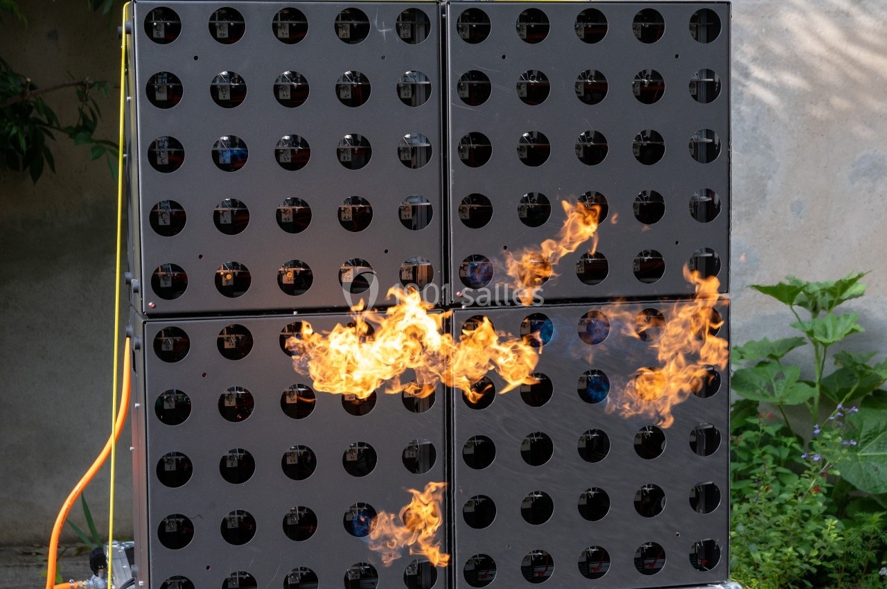 Panneau métallique perforé avec des flammes visibles au centre, installé en extérieur près d'un mur et de végétation.