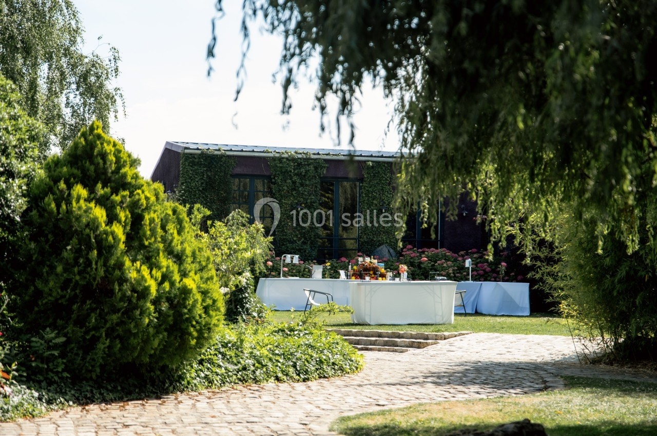 Table dressée en extérieur dans un jardin verdoyant, entourée de buissons et d'arbres, sous un ciel dégagé.