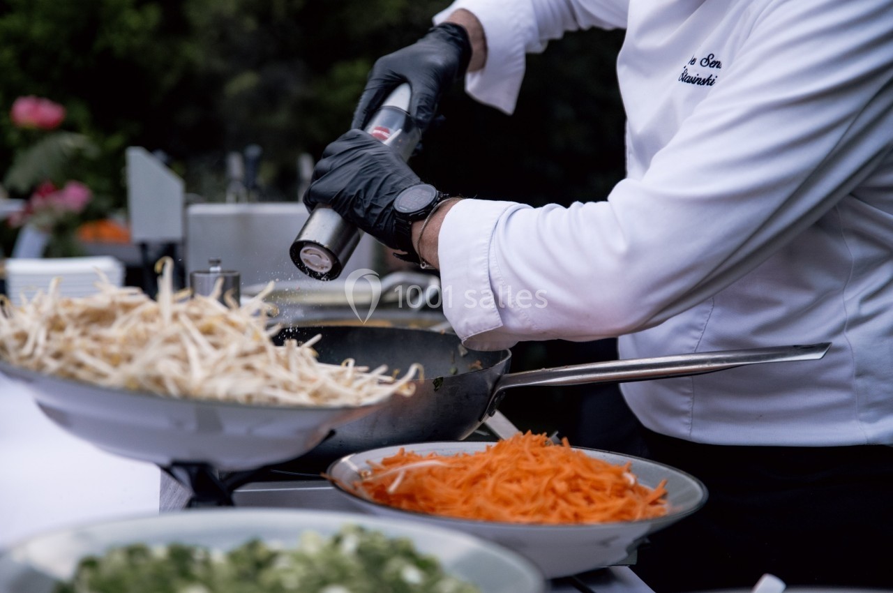 Un chef en tenue blanche assaisonne un plat dans une poêle, entouré de bols de légumes préparés.