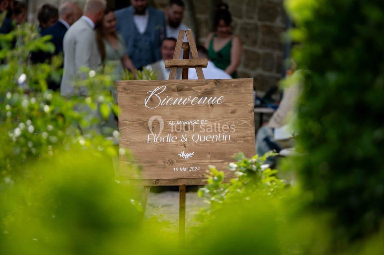 Panneau en bois indiquant ’Bienvenue au mariage de Élodie & Quentin, 18 mai 2024’, entouré de verdure.