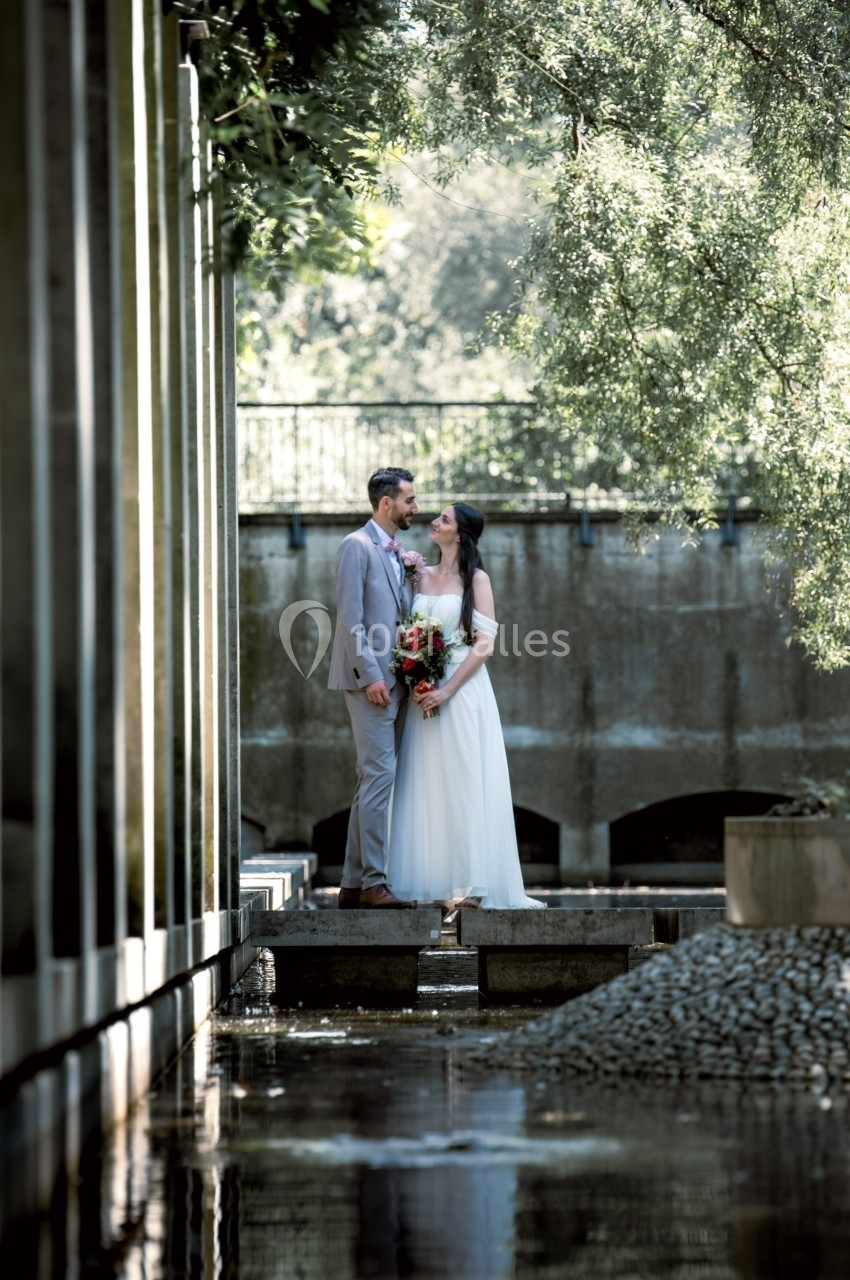 Un couple en tenue de mariage se tient sur une passerelle au-dessus de l'eau, entouré de verdure et d'ombres douces.