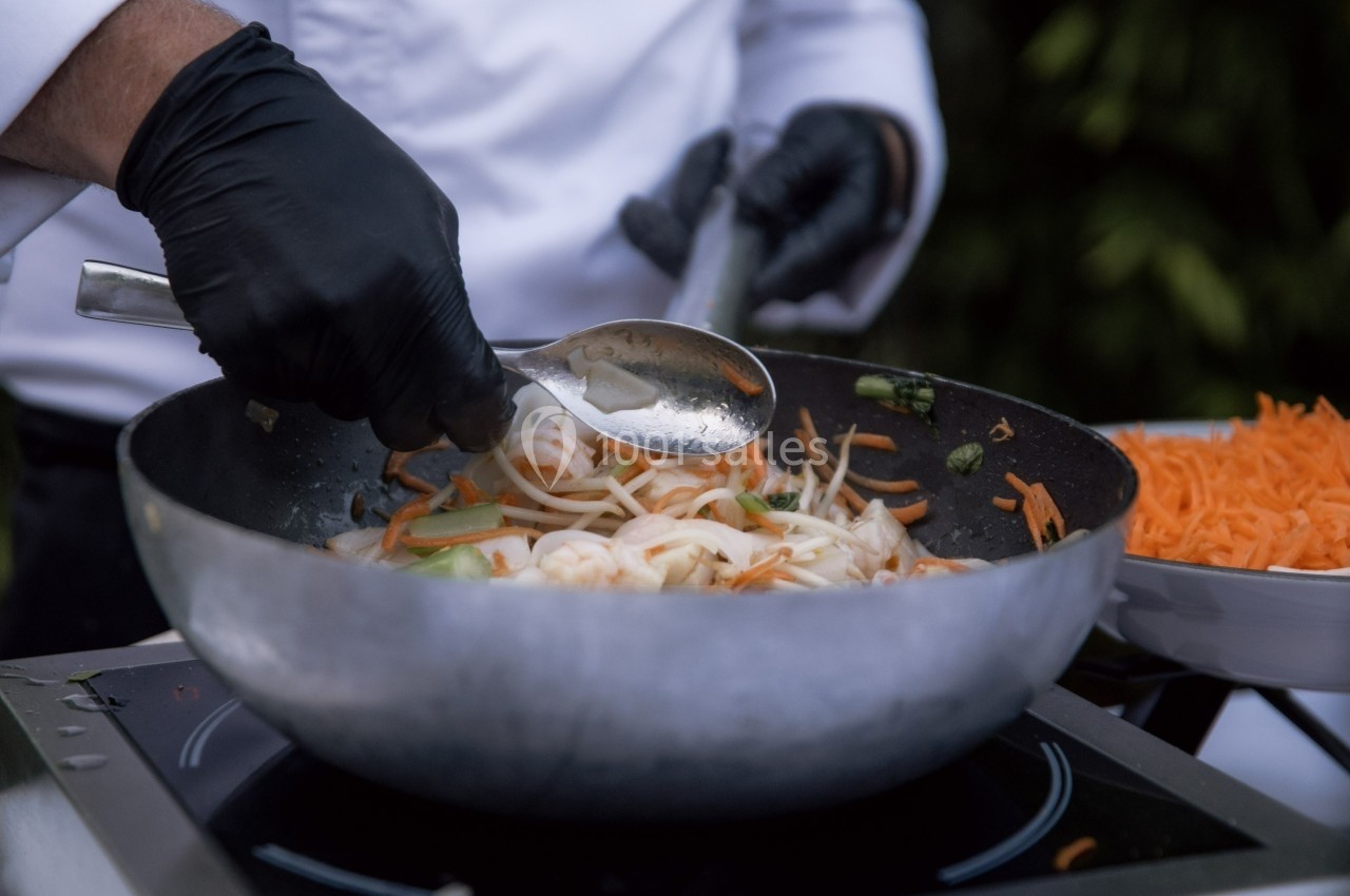 Un chef mélange des légumes sautés dans un wok avec une cuillère en métal, portant des gants noirs.