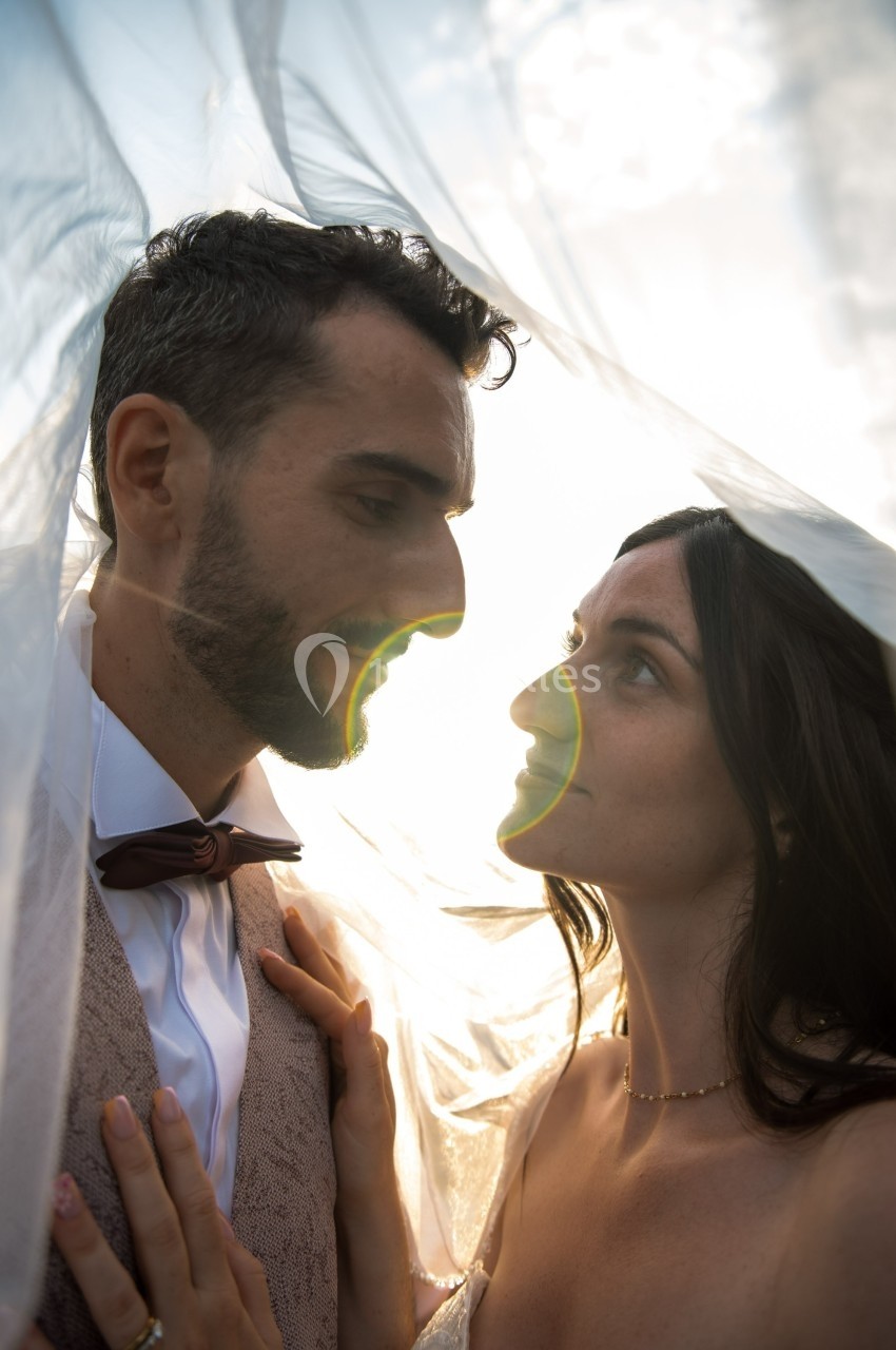 Un couple habillé élégamment se regarde sous un voile translucide, éclairé par une lumière douce.