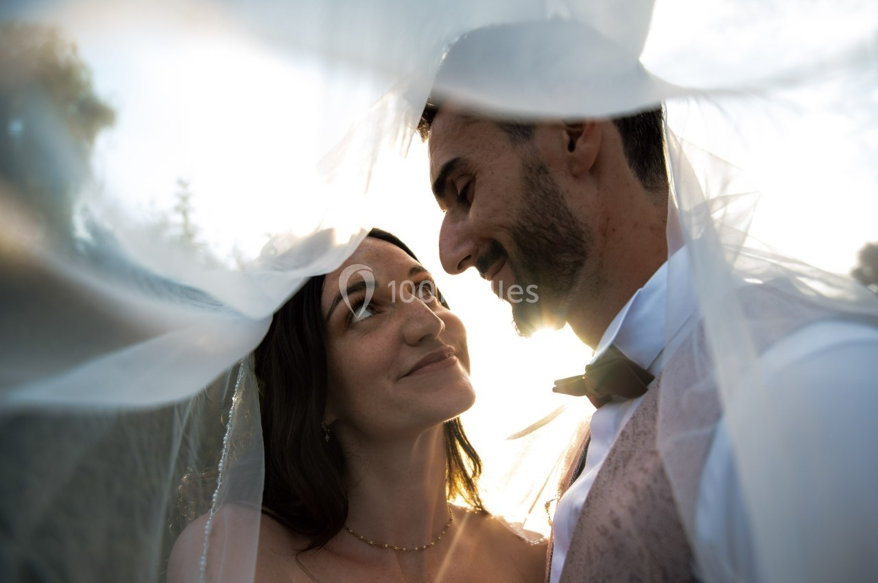 Un couple souriant sous un voile blanc, éclairé par une lumière douce en extérieur.