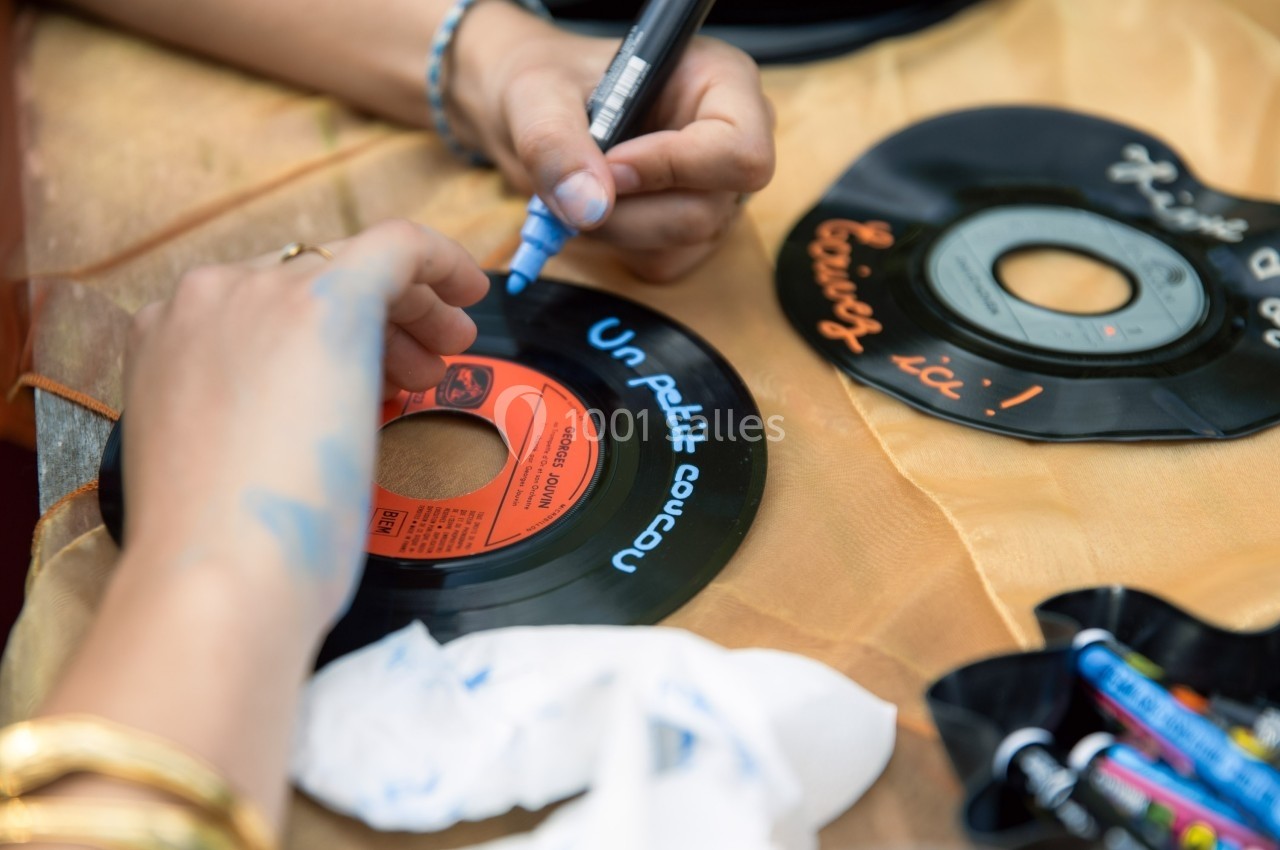 Deux mains personnalisent des disques vinyles avec des marqueurs colorés sur une table recouverte de tissu beige.