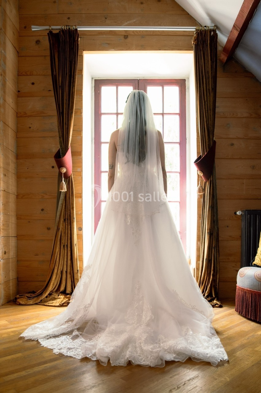 Une mariée en robe blanche vue de dos, debout devant une fenêtre encadrée de rideaux dans une pièce en bois.
