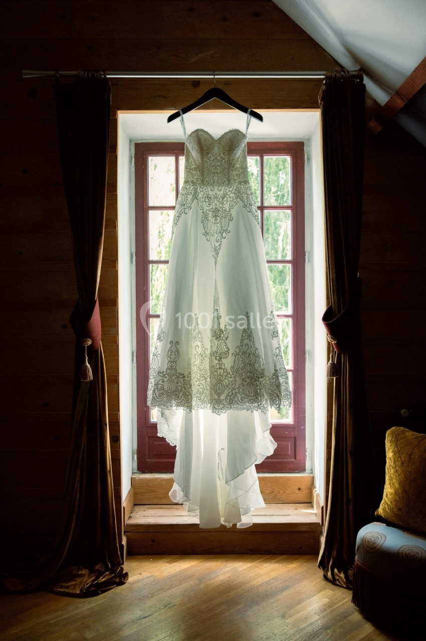 Robe de mariée blanche avec dentelle suspendue devant une fenêtre encadrée de rideaux dans une pièce en bois.
