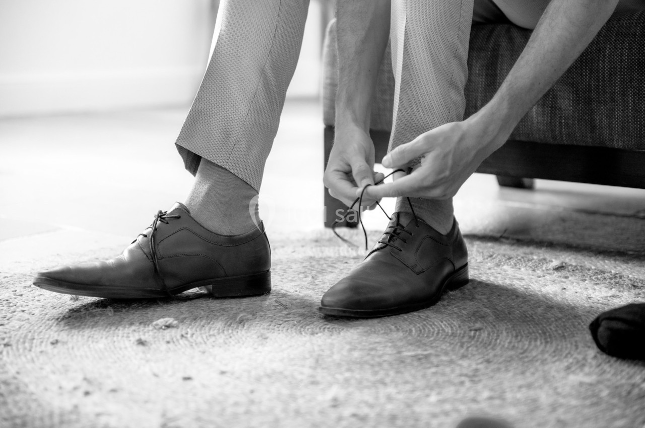 Un homme noue les lacets de ses chaussures habillé d'un pantalon clair, assis sur le bord d'un canapé.
