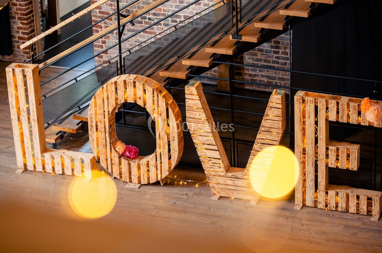 Lettres géantes en bois formant le mot ’LOVE’, décorées de lumières et fleurs, devant un escalier en métal.