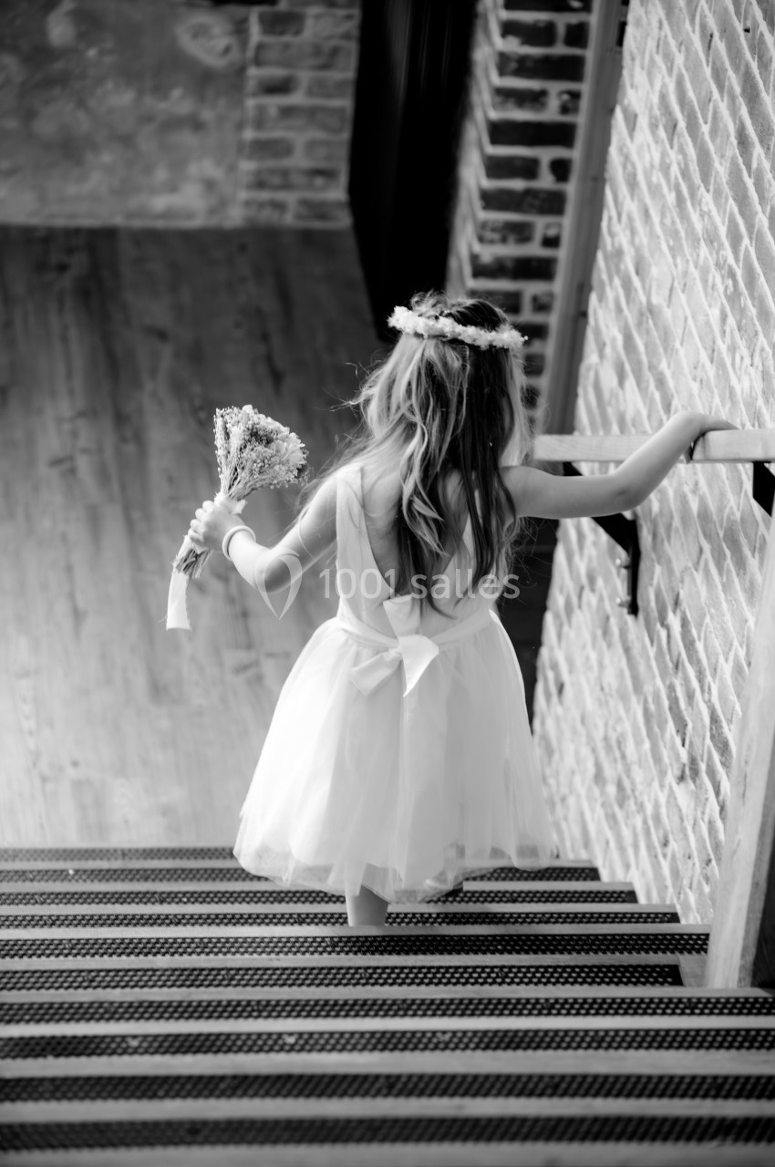 Une fillette en robe blanche et couronne de fleurs monte un escalier en tenant un bouquet.