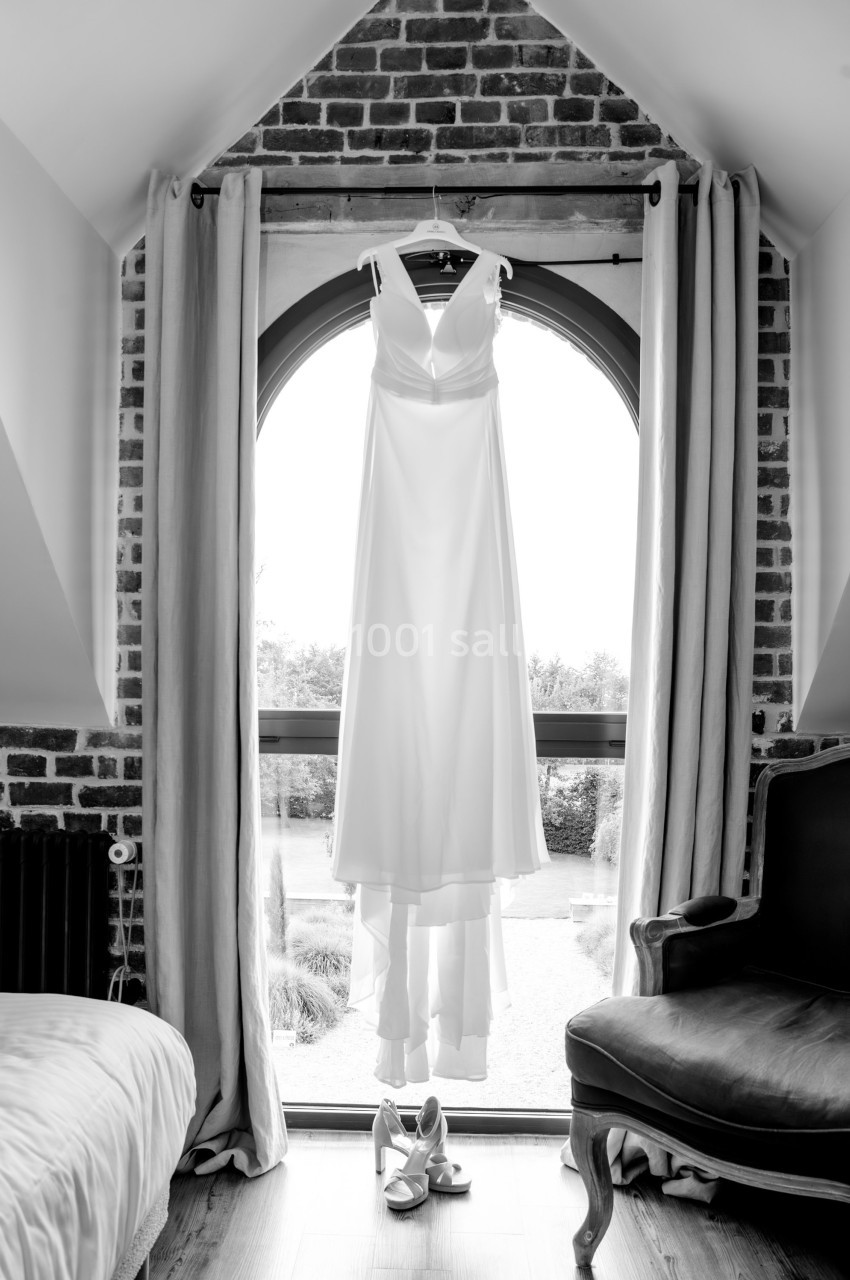 Robe de mariée blanche suspendue devant une fenêtre en arche, avec des chaussures posées au sol.