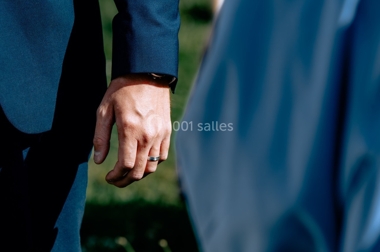 Main d'une personne portant une alliance, vêtue d'un costume bleu, avec un arrière-plan flou de verdure.