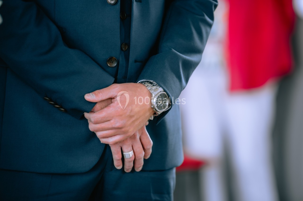 Un homme en costume bleu, mains croisées devant lui, portant une montre et une bague argentées.