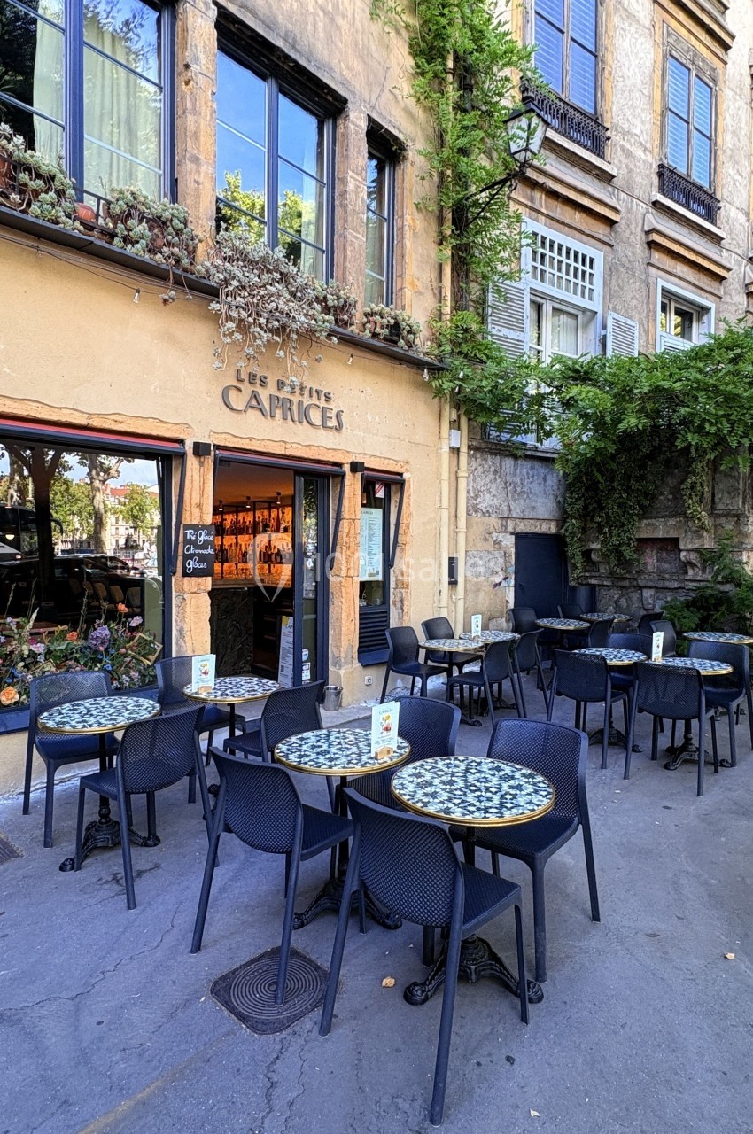 Terrasse d'un café avec des tables et chaises noires, située devant une façade en pierre ornée de plantes.