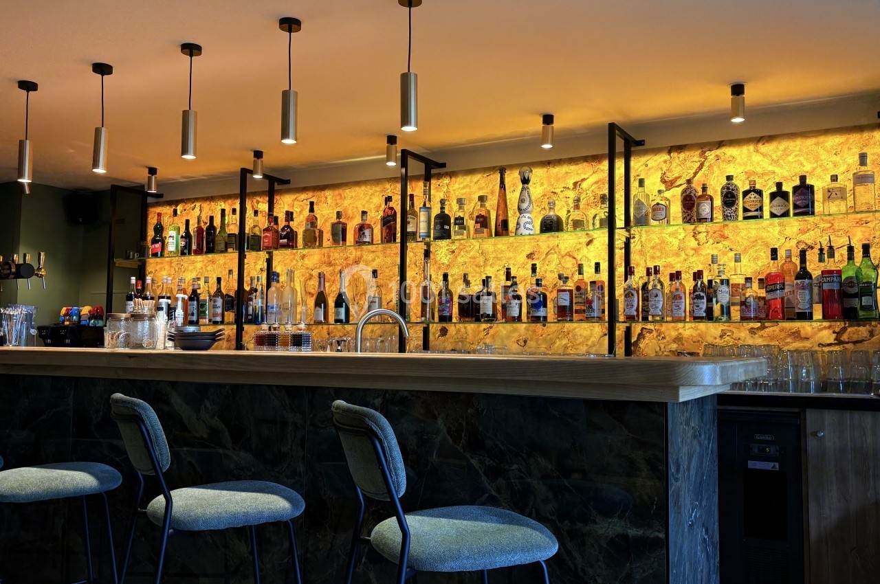 Bar moderne avec comptoir en bois, tabourets rembourrés et étagères illuminées présentant des bouteilles d'alcool.