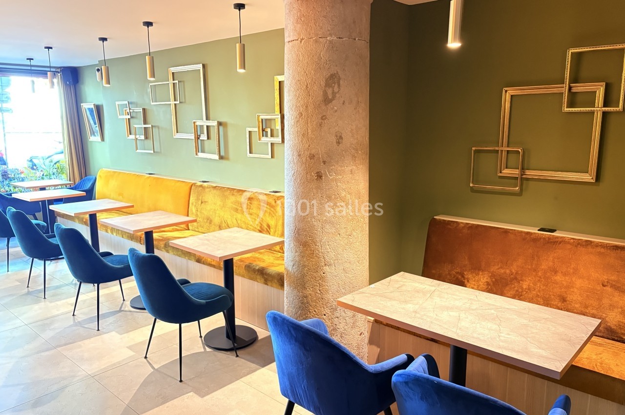 Salle de restaurant moderne avec murs verts, banquettes orange, chaises bleues et tables en bois clair.