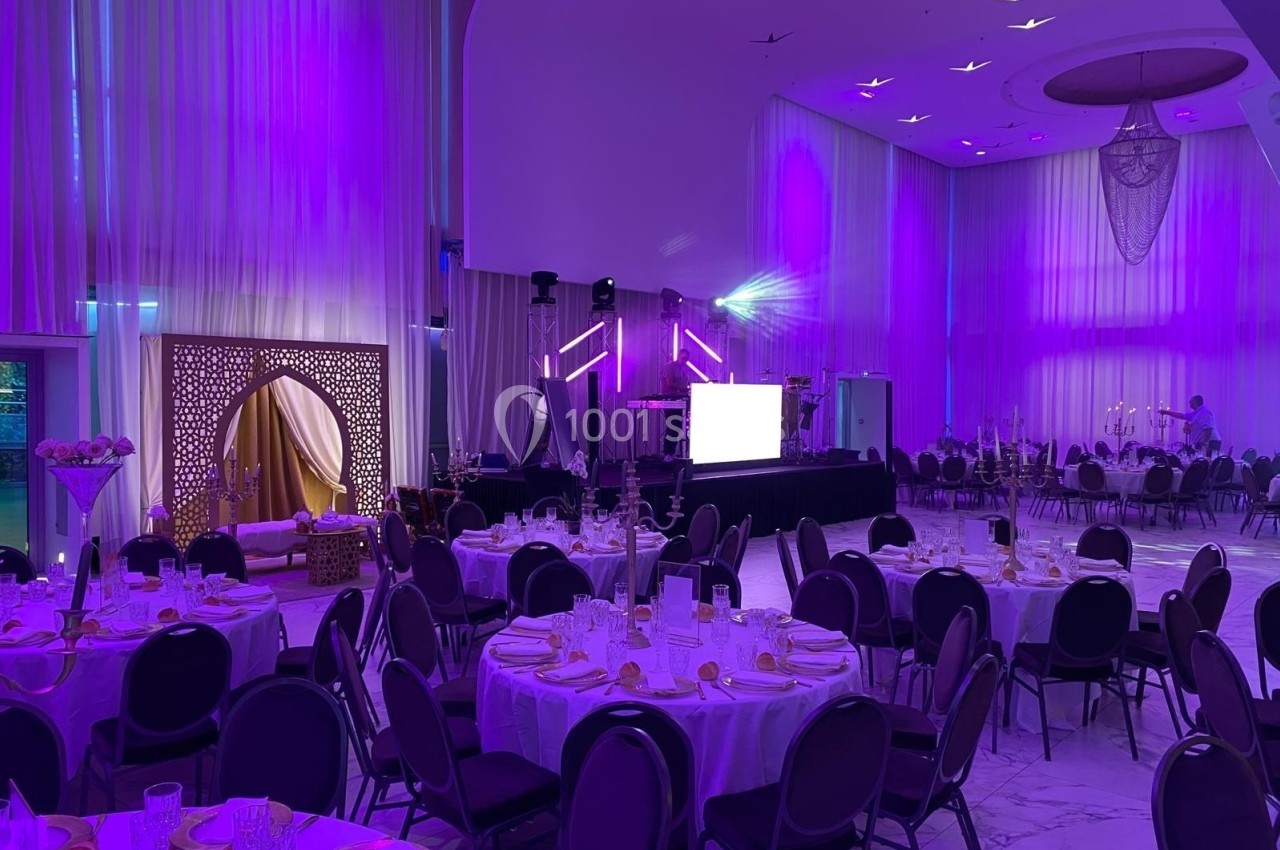Salle de réception décorée avec des tables rondes dressées, éclairage violet et espace scénique en arrière-plan.