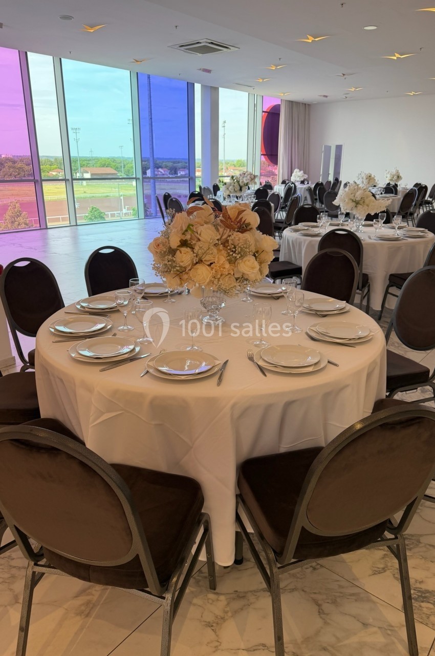 Salle de réception lumineuse avec tables rondes dressées, nappes blanches et centre de table floral, vue extérieure colorée.