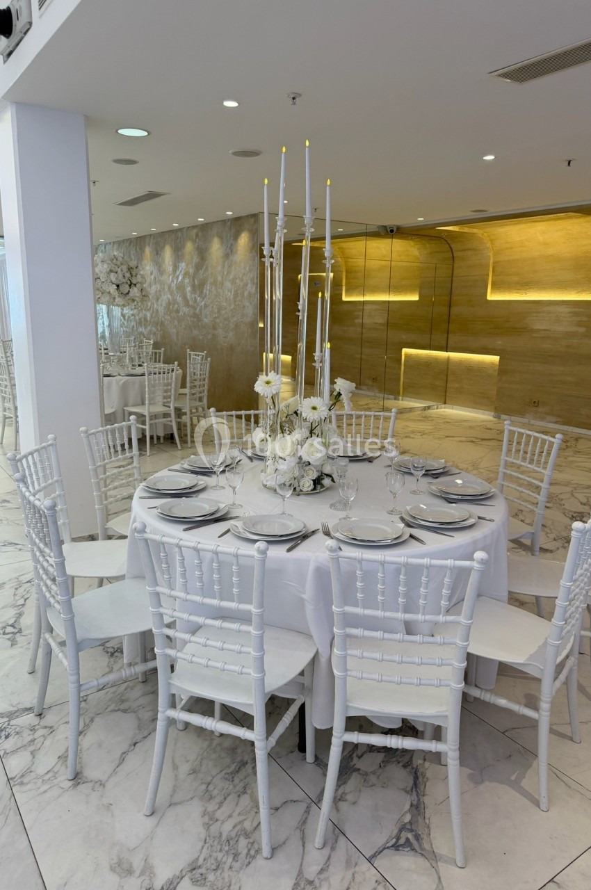 Table ronde élégamment dressée avec des chaises blanches, décorée de bougies et de fleurs blanches dans une salle lumineuse.
