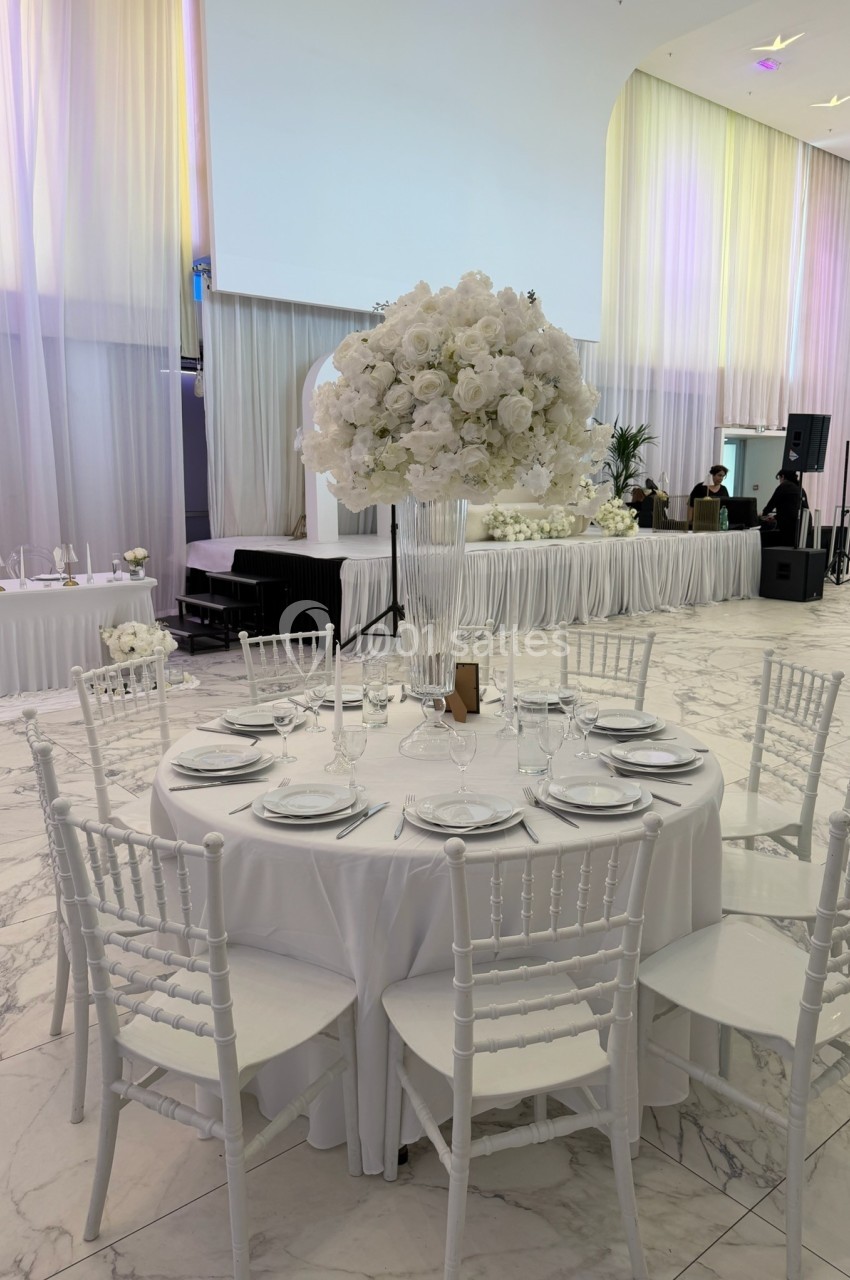 Table ronde décorée pour un événement, avec nappes blanches, chaises assorties et un centre de table floral blanc.