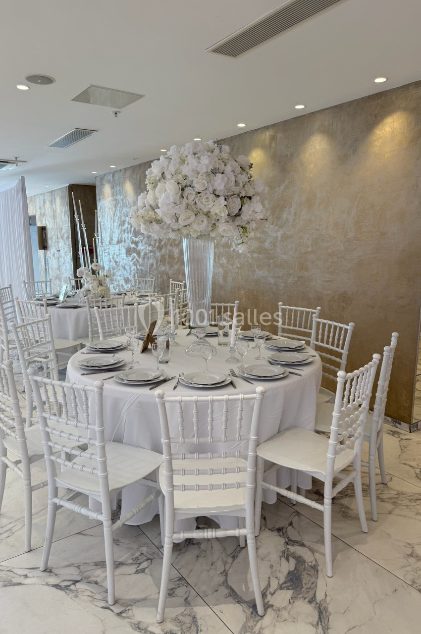 Table ronde élégamment dressée avec nappes blanches, chaises assorties et centre de table floral blanc dans une salle…