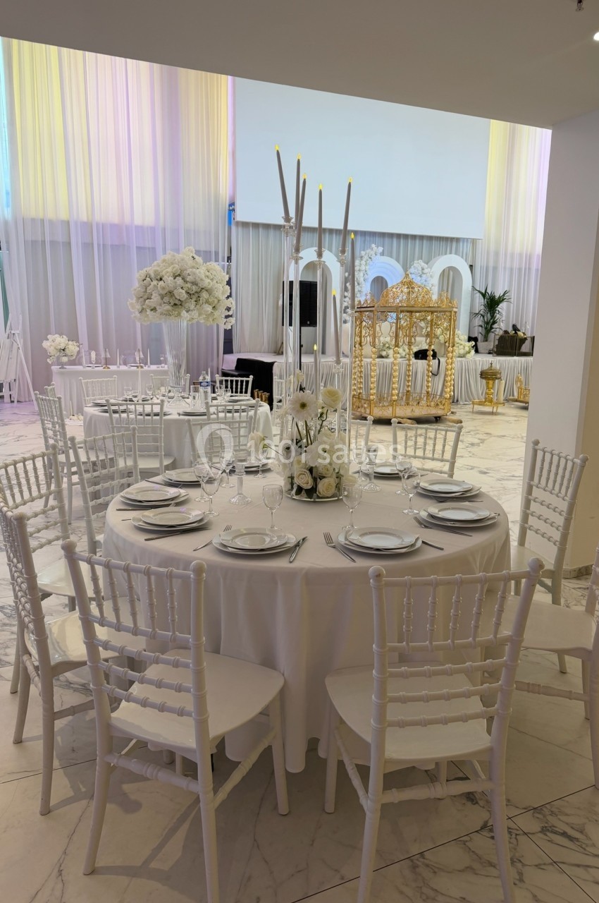 Salle de réception élégante avec tables rondes dressées, chaises blanches et décoration florale blanche.