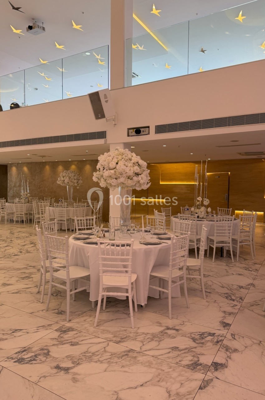 Salle de réception lumineuse avec tables rondes dressées, chaises blanches et décorations florales élégantes.