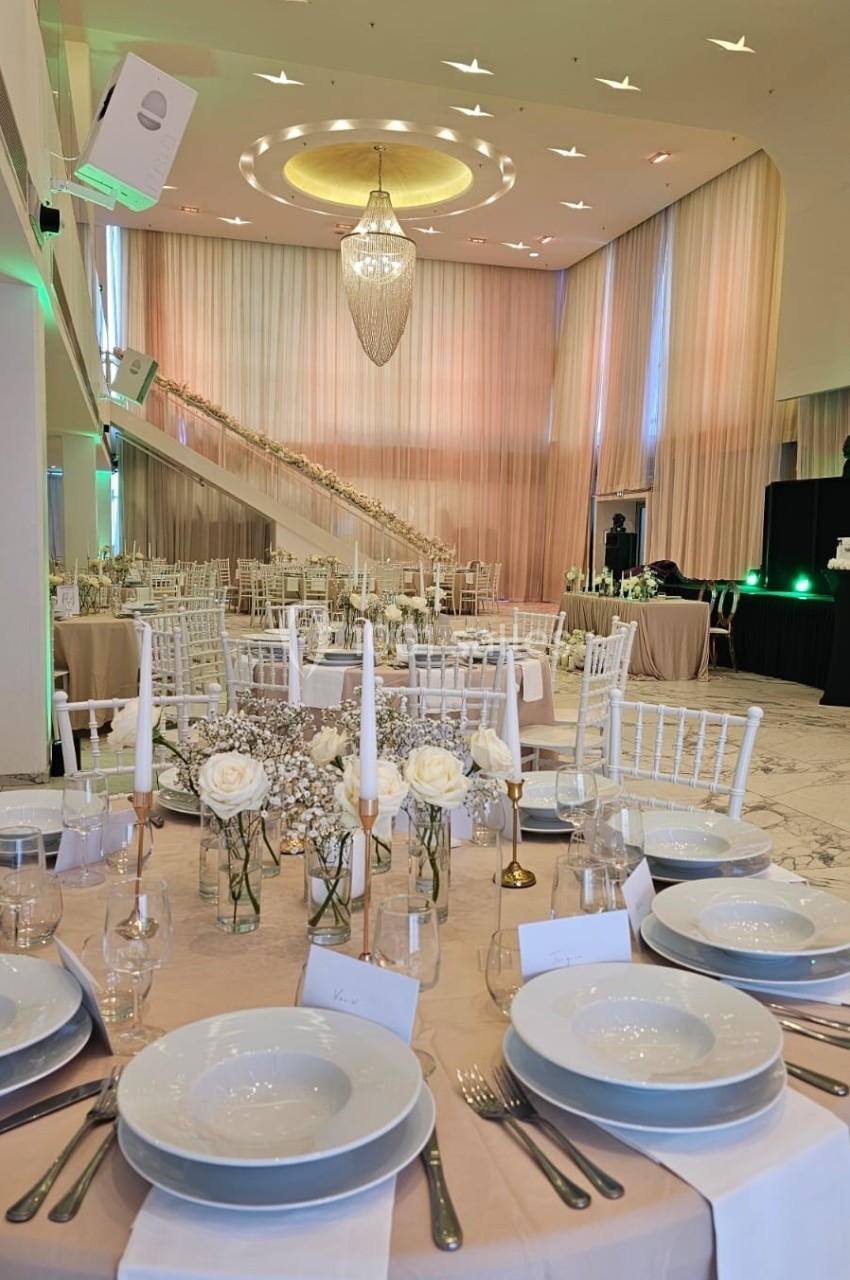 Salle de réception élégante avec tables dressées, chandeliers, roses blanches et escalier décoré en arrière-plan.