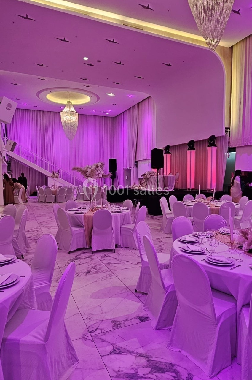 Salle de réception élégante avec tables rondes dressées, chaises blanches et éclairage rose-violet.