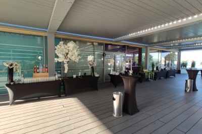 Salle extérieure aménagée avec des tables hautes noires, décorations florales blanches et lumière naturelle.