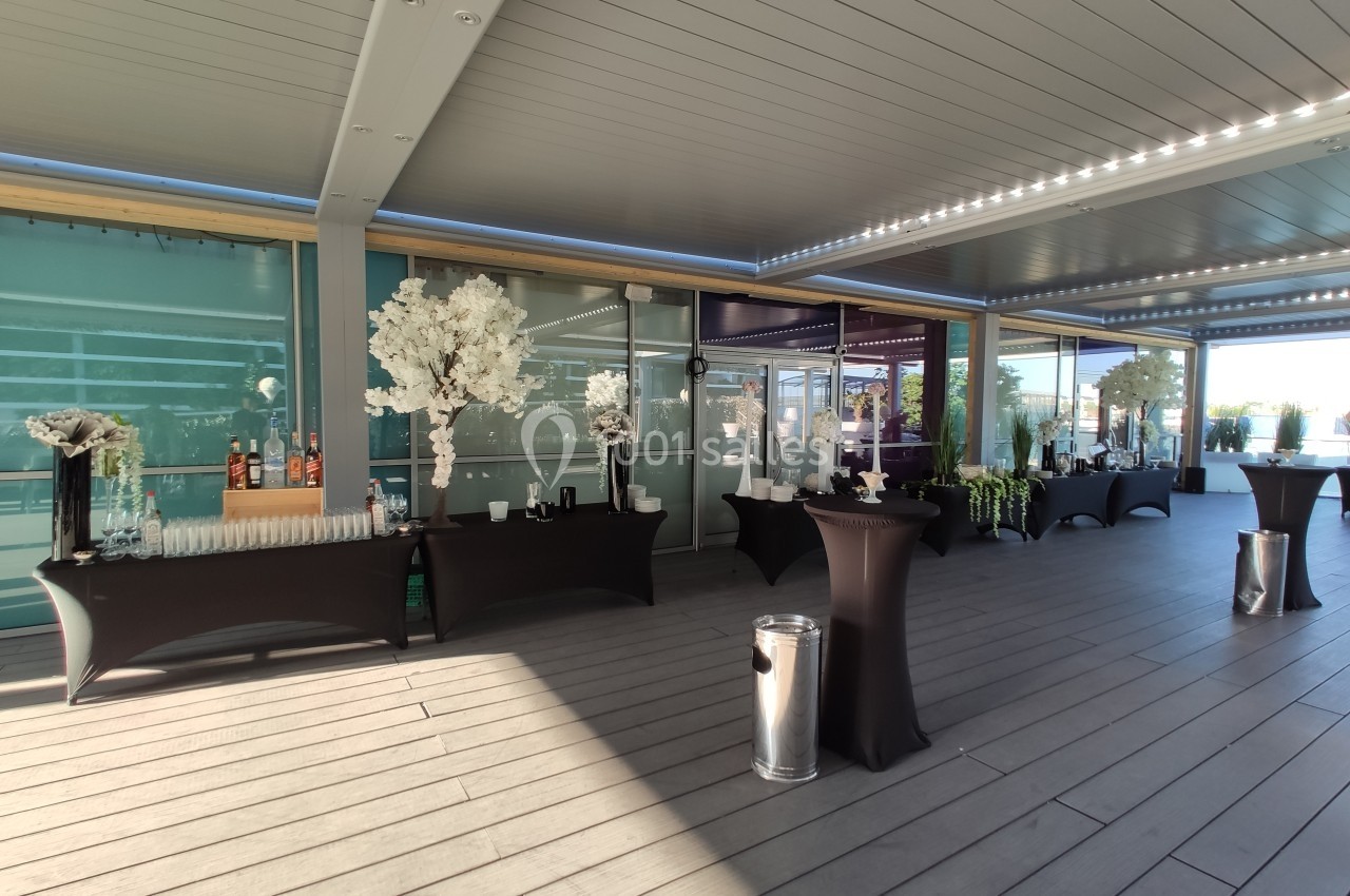 Salle extérieure aménagée avec des tables hautes noires, décorations florales blanches et lumière naturelle.