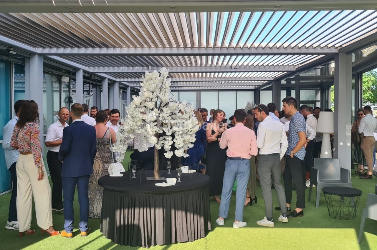 Groupe de personnes discutant debout sous une pergola moderne avec un décor floral au centre.