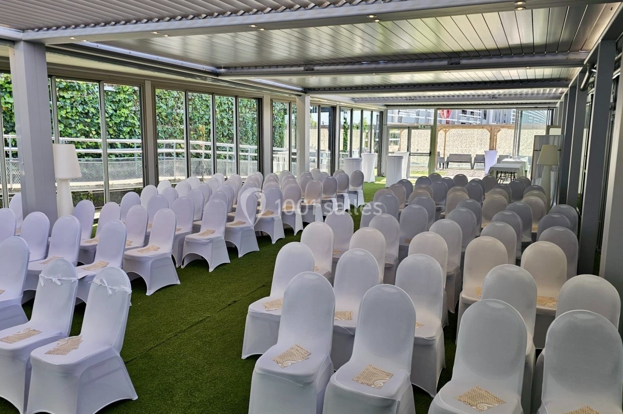 Salle de réception avec des rangées de chaises blanches alignées sous une structure couverte, sur un sol en gazon artificiel.